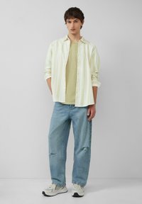 Gestreiftes, hellgelbes und weißes Hemd über einem beigen T-Shirt, kombiniert mit hellblauen, verwaschenen Jeans und grauen Sneakers.