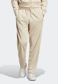 Béžové tepláky s vzorem listů, elastický pas a šňůrka na stažení. Materiál vypadá měkký a pohodlný. Nošeny s bílými teniskami.