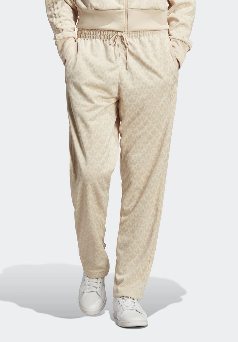 Béžové tepláky s vzorem listů, elastický pas a šňůrka na stažení. Materiál vypadá měkký a pohodlný. Nošeny s bílými teniskami.