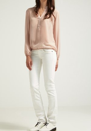 Lichtroze blouse met lange mouwen, V-hals en knoopdetails, gecombineerd met witte slim-fit jeans en zwarte veterschoenen.