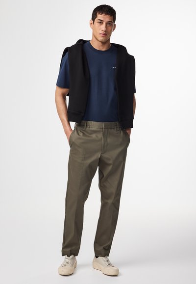 Marine t-shirt, zwarte hoodie over de schouders, olijfgroene broeken en lichtgrijze sneakers. Soepele stof en een ontspannen pasvorm.