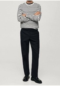 Camicia a maniche lunghe bianca e blu a righe con scollatura rotonda, abbinata a pantaloni neri a sigaretta e scarpe nere, in piedi con le braccia incrociate.