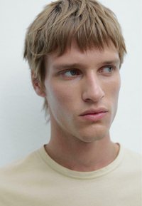 Ingrandimento del viso di un modello con capelli castano chiaro e occhi blu, che indossa una camicia beige. La pelle ha una texture liscia con dettagli sottili.