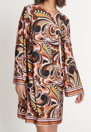 Kobieta ubrana w sukienkę z długim rękawem o wirującym, wielokolorowym abstrakcyjnym wzorze w odcieniach brązu, musztardy, różu, czerni i bieli.
