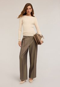 Kobieta w kremowej, prążkowanej bluzce z długim rękawem, brązowych, szerokich spodniach w paski, beżowych szpilkach, trzymająca torebkę w kolorze taupe na neutralnym tle.