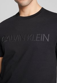 Černé tričko s kulatým výstřihem vyrobené z měkké bavlny, s embosovaným logem "CALVIN KLEIN" na přední straně v tónovém provedení. Krátké rukávy, klasický střih.