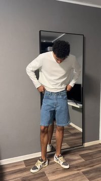 Άνδρας με λευκό μακρυμάνικο πουκάμισο, μπλε τζιν σορτς και αθλητικά παπούτσια ρυθμίζει τα σορτς του ενώ στέκεται μπροστά σε έναν καθρέφτη ολόσωμο σε ένα δωμάτιο.