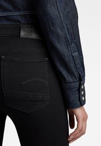 Camisa de denim oscuro con una chaqueta negra ajustada, que presenta botones a presión metálicos y costuras de bolsillo visibles en los pantalones negros.