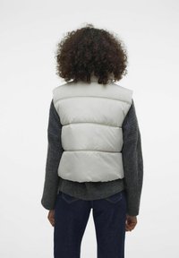 Gilet imbottito grigio chiaro con colletto alto, caratterizzato da cuciture orizzontali e un design imbottito. Indossato sopra un maglione scuro e jeans.