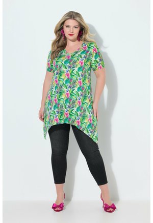 Kobieta w zielonej kwiecistej tunice, czarnych legginsach, różowych kolczykach i różowych butach na obcasie z kokardą stoi na białym tle.