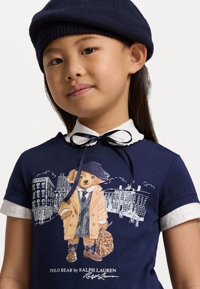 Μπλούζα από βαμβάκι σε σκούρο μπλε χρώμα με άσπρο γιακά, διαθέτοντας γραφικό με μια αρκούδα σε παλτό και καπέλο, που κρατάει μια τσάντα, μπροστά από φόντο με αστική τοπογραφία.