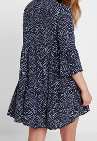 Granatowa sukienka w białe abstrakcyjne kropki, z rozkloszowanymi rękawami i warstwowym krojem spódnicy, prezentująca miękki materiał i luźny krój.