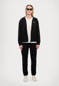 Felpa con zip nera sopra una maglietta bianca, abbinata a pantaloni della tuta neri e sneakers bianche. Presenta un dettaglio di marca sulla felpa.