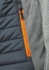 Szary, teksturowany materiał z ciemnoszarym, pikowanym fragmentem. Pomarańczowy zamek błyskawiczny po lewej stronie dodaje kontrastu i detali.