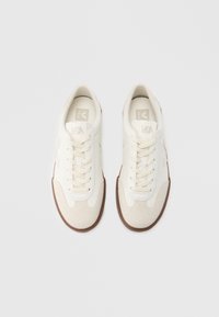 Bijele tenisice s vezicama izrađene od glatke kože i brušene kože, s bež kapicom na prstu, smeđim gumenim potplatom i suptilnim otiskom logotipa na jeziku.