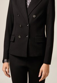 Czarny dwurzedowy żakiet z błyszczącymi metalowymi guzikami, kieszeniami bocznymi i teksturowanym materiałem. Slim-fit design w połączeniu z dopasowanymi czarnymi spodniami.