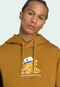 Brun hættetrøje lavet af bomuld med et grafisk motiv af en leopard iført en kasket og teksten "CASUAL SPORTS CLUB adidas" på fronten.