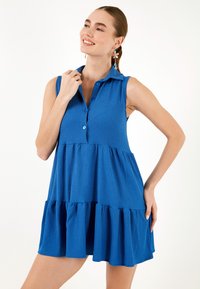 Rochie albastră fără mâneci, cu guler cu nasturi, tiv cu volane în straturi și țesătură texturată. Prezintă o croială lejeră și o culoare vibrantă.