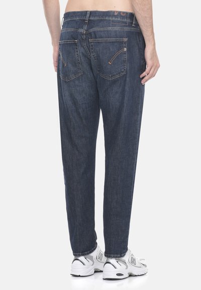 Persona che indossa jeans in denim blu scuro e sneaker bianche New Balance, mostrata da dietro su uno sfondo bianco.