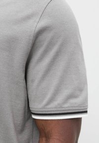 Korte grijze polo met mouwen, afgewerkt met wit en zwart gestreepte rand op een donkerhuidige arm tegen een effen achtergrond.