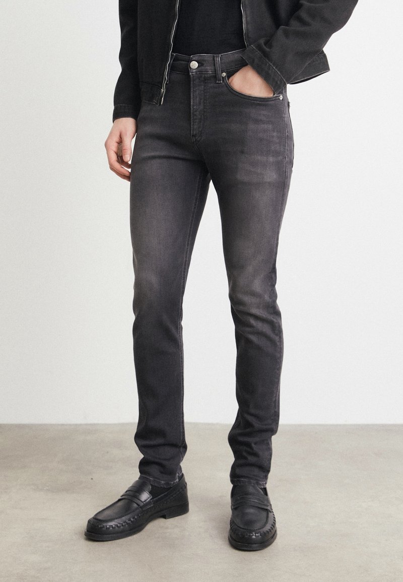Jean ajustado negro de denim, con cinco bolsillos, cremallera en la bragueta y un leve desgaste. Combinados con mocasines negros.