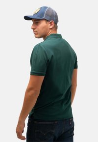 Grön pikétröja med korta ärmar, kombinerad med en blå meshkeps med en rund lapp, sedd från sidan tillsammans med jeans.