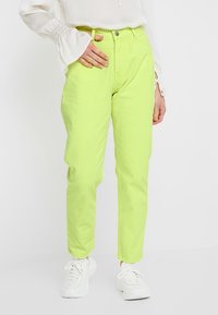 Pantalones de algodón de un vibrante verde lima con corte recto, que incluyen un cierre de botón y cremallera, además de bolsillos laterales. Llevados con zapatillas blancas.