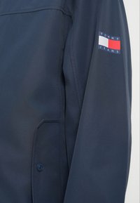 Ναυτικό μπλε μπουφάν με απαλή υφή, διαθέτει τσέπη και μικρή ετικέτα λογότυπου "Tommy Jeans" με κόκκινες, λευκές και μπλε λεπτομέρειες στον μανίκι.