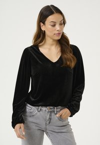 Schwarze Samt-Bluse mit langen Ärmeln und V-Ausschnitt, ausgestattet mit gerafften Details an den Schultern und einer weichen, glatten Textur. Getragen mit hellgrauen Jeans.