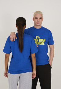 Tricourile de bumbac bleu au un slogan galben, "1% MAI BINE DECÂT IERI." O persoană poartă pantaloni de trening gri, în timp ce cealaltă poartă pantaloni negri.