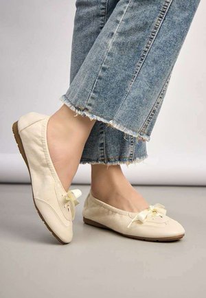 Pieds portant des ballerines couleur crème avec des nœuds en ruban, associées à un jean bleu court à ourlet effiloché.