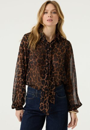 Frau schaut nach vorne, trägt eine transparente, braune Bluse mit Leopardenmuster und Halsbindedetail sowie dunkelblaue Jeans, steht vor einem schlichten Hintergrund.