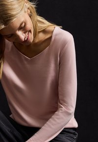 Helles rosa Langarmoberteil mit V-Ausschnitt, weichem Material, tailliertem Design und dezenten Nähdetails, kombiniert mit schwarzen Hosen.