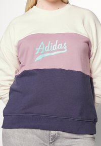 Sudadera con bloques de color morado, crema y azul marino hecha de tela suave. Presenta un logo bordado de "Adidas" en azul claro en el pecho.