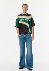 Grafische t-shirt met een groene en bruine gradient, met de tekst "Echoes of Glory". Gecombineerd met een wijde blauwe jeans en zwarte schoenen.