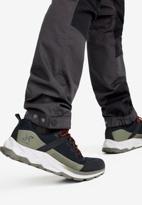 Person som går och bär svarta och olivgröna vandringsskor med vita sulor och mörkgrå byxor med tryckknappar vid anklarna.