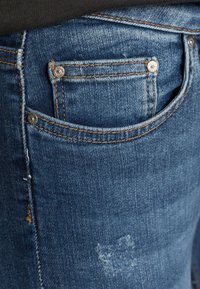 Jeans de mezclilla en un color azul intermedio con ligero desgaste, costuras de cobre y un pequeño bolsillo frontal con remaches de metal.