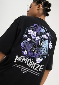 Junge Person mit geflochtenem Haar, die eine Brille und ein schwarzes T-Shirt trägt, auf dem auf der Rückseite lila Koi-Fische, Blumen und stilisierte Schrift zu sehen sind.