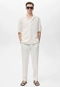 Camisa blanca de manga corta de punto texturizado con cuello, combinada con pantalones de color claro y sandalias negras, que presenta un aspecto limpio y moderno.