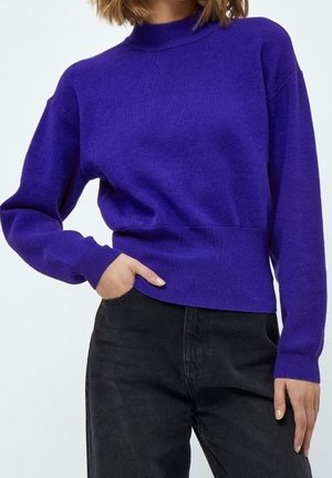 Kvinde iført en lilla langærmet sweater med ribkanter ved ærmeender og taljebånd, kombineret med sorte høj-taljebukser, hånd i lommen.