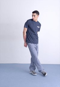 T-shirt en coton bleu marine avec un logo sur la poitrine, associé à un pantalon gris clair et des chaussures de sport grises, sur un fond uni.