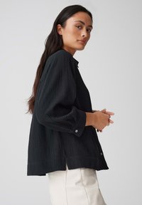 Zwarte, getextureerde shirt met een knoopsluiting bij de kraag en lange mouwen, met een losse pasvorm en zijsplitten. gedragen over lichte broek.