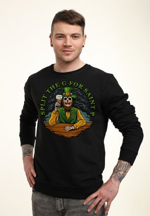 Mand med kort brunt hår og tatoveringer iført en sort sweatshirt med en farverig skelet-leprechaun, der holder en drink.
