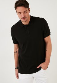 Polo negro de manga corta con textura acanalada, que presenta una tapeta de botones y un pequeño logotipo bordado en el pecho.
