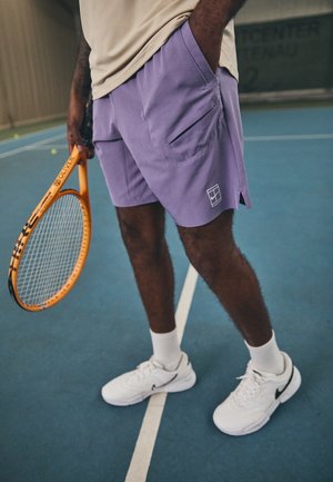 Osoba stojąca na krytym korcie tenisowym, trzymająca pomarańczową rakietę tenisową, ubrana w fioletowe szorty, beżową koszulkę, białe skarpetki i białe buty Nike.