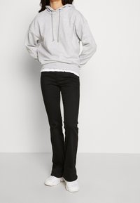 Světle šedá mikina s kapucí a malým logem, kombinovaná s černými rozšířenými džíny a bílými teniskami. Oblečení má neformální design.