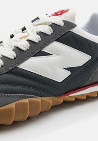 Zapatilla gris con detalles en blanco, materiales de tela y cuero, parte superior texturizada, suela de goma con surcos y cierre con cordones.