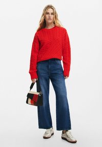 Maglione rosso a coste, jeans blu a gamba larga e una borsa a tracolla a motivi neri, rossi e crema. L'outfit presenta materiali texturizzati e un design informale.
