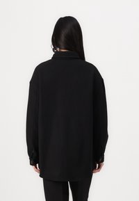 Svart oversized skjorta med krage, långa ärmar och knappade ärmslut. Tillverkad av ett mjukt material med en slät textur. Bakvy.