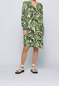 Grünes und weißes langärmliges Kleid mit großem Blattmuster, V-Ausschnitt und gerafften Bündchen. Kombiniert mit weißen Sandalen und einer dicken schwarzen Sohle.